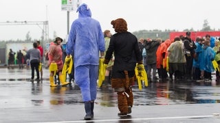 Zwei Festivalbesucher laufen über nassen Asphalt über das Gelände von Rock am Ring. (Foto: IMAGO, picture-alliance / Reportdienste, EIBNER/Daniel Reinelt) Zwei Festivalbesucher laufen über nassen Asphalt über das Gelände von Rock am Ring.