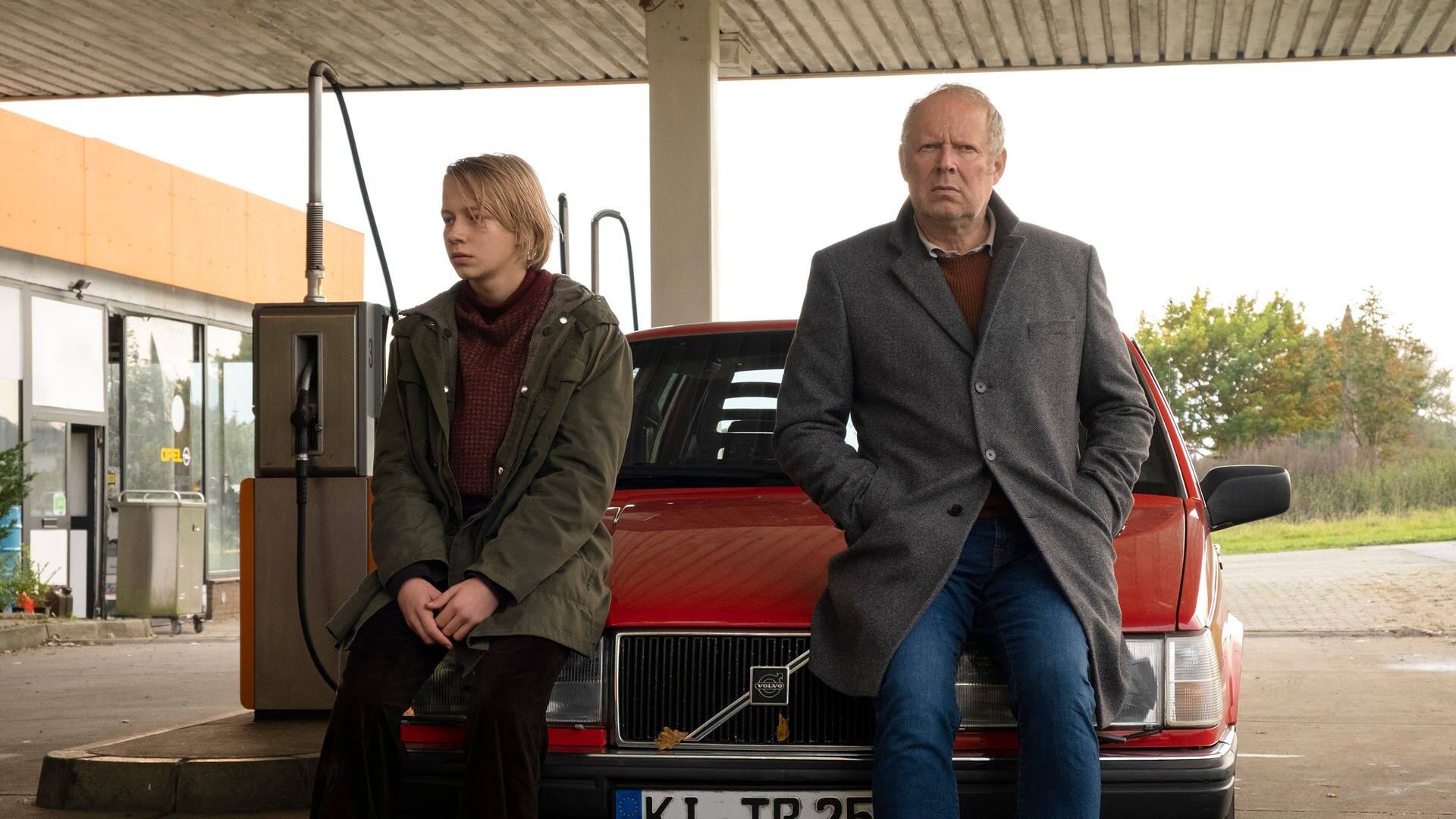 Borowski und sein jüngeres Ich sitzen auf der Motorhaube eines roten Autos an einer Tankstelle. (Foto: NDR/Christine Schroeder)