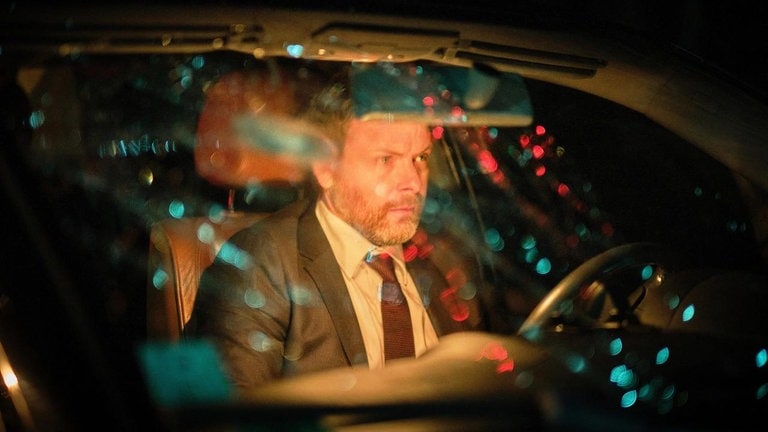 Foto: SWR, SWR / Benoît Linder Der Täter sitzt nach der Tat im Auto, der Blick geht durch die Windschutzscheibe, Lichter spiegeln sich. (Foto: SWR, SWR / Benoît Linder)