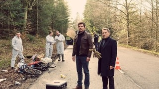 Tatort-Kommissare schauen sich am Tatort Landstrasse um. Ein Fahrrad des Opfers daneben.