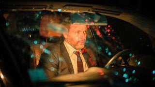 Der Täter sitzt nach der Tat im Auto, der Blick geht durch die Windschutzscheibe, Lichter spiegeln sich.
