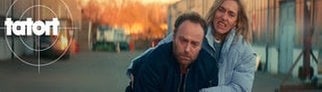 Kommissar Karow aus dem Tatort wird von einer Frau gestützt. Beide verziehen vor Schmerz das Gesicht, als sie in Berlin im Sonnenuntergang durch eine Strasse laufen.