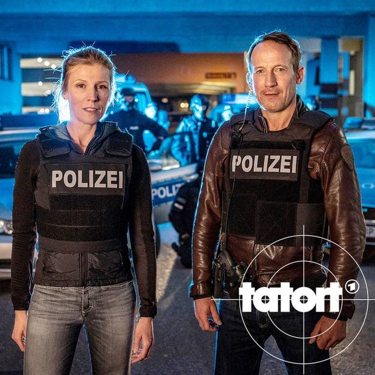 Julia Grosz und Thorsten Falke stehen in Einsatzkleidung nachts vor Polizeiautos und dem SEK