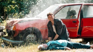 Tatort-Kritik Saarbrücken „Der Fluch des Geldes“: Kommissar Hölzer kniet neben einem verunfallten Auto neben einer alten Frau