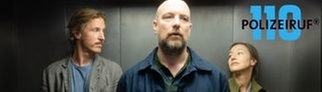 Polizeiruf „Funkensommer“ München: In einem Aufzug stehen hinten Chris und Dennis und lächeln sich an. Im Vordergrund steht Eden und schaut genervt