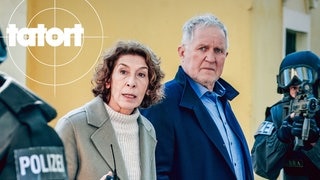 Szenenbild aus dem Tatort „Wir sind nicht zu fassen!“ aus Wien: Die Ermittler Moritz Eisner und Bibi Fellner zwischen zwei bewaffneten Polizisten
