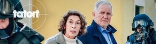 Szenenbild aus dem Tatort „Wir sind nicht zu fassen!“ aus Wien: Die Ermittler Moritz Eisner und Bibi Fellner zwischen zwei bewaffneten Polizisten