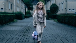 Szenenbild aus dem Tatort „Feuer“ aus Dortmund: Kleines Mädchen, barfuß und rußverschmiert, steht allein mit Teddybär auf gepflasterter Straße (Foto: ard-foto s2-intern/extern, WDR/Martin Rottenkolber) Szenenbild aus dem Tatort „Feuer“ aus Dortmund: Kleines Mädchen, barfuß und rußverschmiert, steht allein mit Teddybär auf gepflasterter Straße