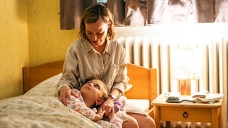 Szenenbild aus dem Tatort „Feuer“ aus Dortmund: Frau sitzt auf dem Bett, hält ihr Kind schützend im Arm. Zimmer wirkt ruhig, warm beleuchtet durch Nachttischlampe. (Foto: ard-foto s2-intern/extern, WDR/Martin Rottenkolber) Szenenbild aus dem Tatort „Feuer“ aus Dortmund: Frau sitzt auf dem Bett, hält ihr Kind schützend im Arm. Zimmer wirkt ruhig, warm beleuchtet durch Nachttischlampe.