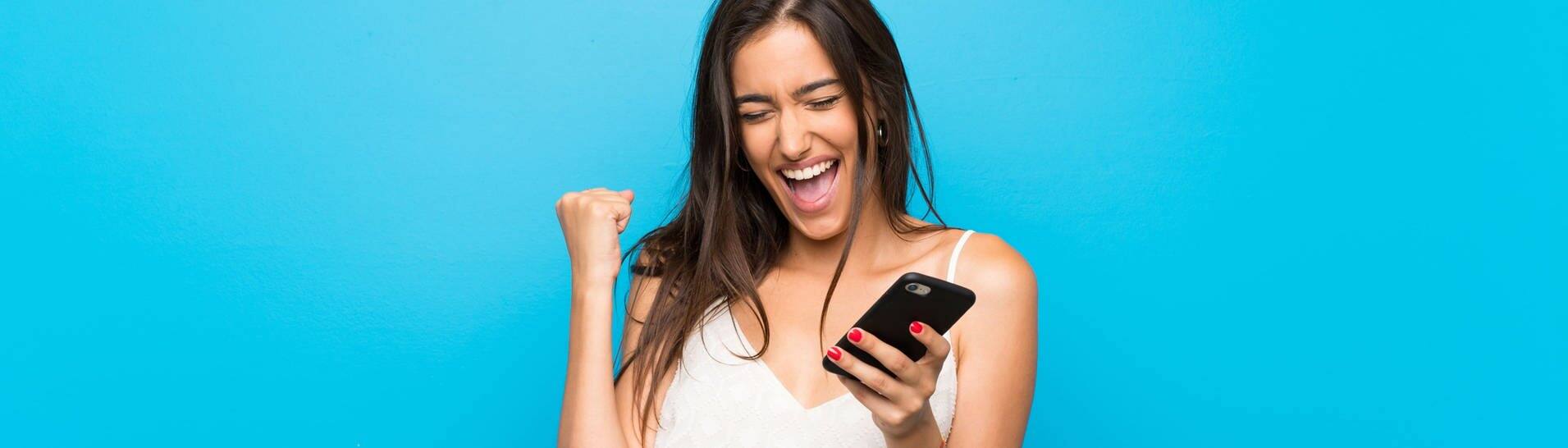 Junge Frau vor blauem Hintergrund mit Handy in der Hand ballt die Faust freudig zusammen und lächelt (Foto: Adobe Stock/luismolinero)
