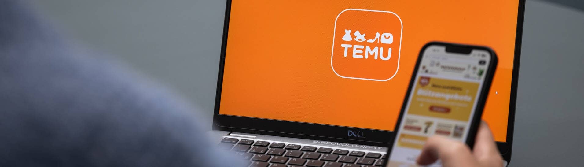 Temu App aus China: Erfahrungsberichte. Person hält Handy mit Temu App vor dem Laptop, auf dem der Onlineshop offen ist