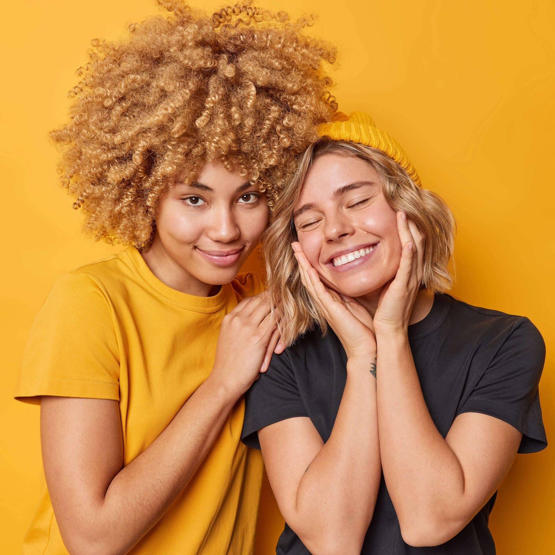 Zwei junge Frauen vor gelbem Hintergrund lächeln, eine hält der anderen die Schulter, die andere schlägt die Hände glücklich übers Gesicht (Foto: Adobe Stock, Adobe Stock / Wayhome Studio)