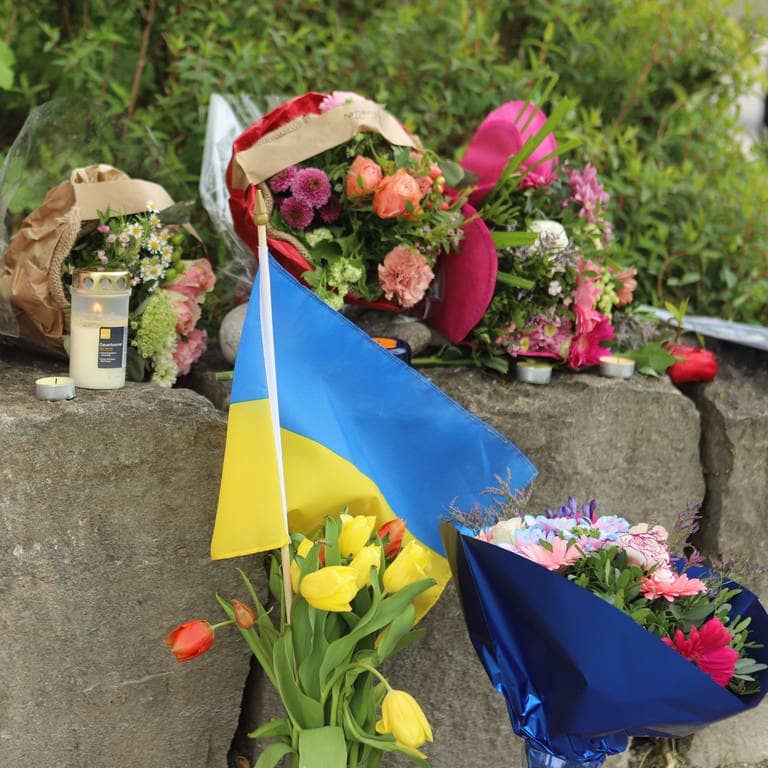 Am Gelände eines Einkaufszentrums in Murnau, an dem am Samstagabend (27.04.2024) zwei Männer aus der Ukraine getötet worden sind, wurden Blumen niedergelegt, sowie eine kleine ukrainische Flagge aufgestellt. Die Polizei konnte einen Mann festnehmen, der als dringend tatverdächtig gilt. Es handelt sich dabei um einen 57 Jahre alten Russen. (Foto: dpa Bildfunk, picture alliance/dpa | Constanze Wilz)