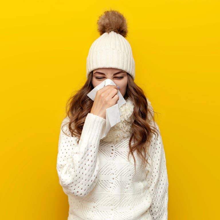 Junge Frau in Winter-Kleidung schnäuzt die Nase. Sie ist vermutlich krank und wird eine Krankschreibung für den Arbeitgeber brauchen oder sich zumindest krankmelden.