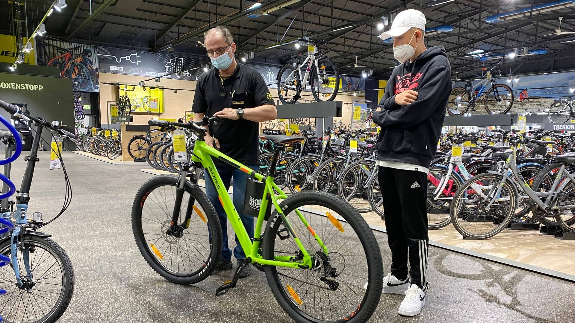 Ein Teenager beim Fahhradkauf in einem Zweirad Fachhandel. Ein Verkauefer beraet den jungen Kunden und stellt den Lenker eines grünen Bikes ein.