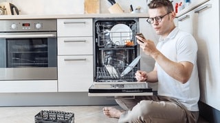 Ein Mann sitzt vor einer Spülmaschine und schaut auf dem Smartphone, was die Spülmaschinen-Symbole bedeuten