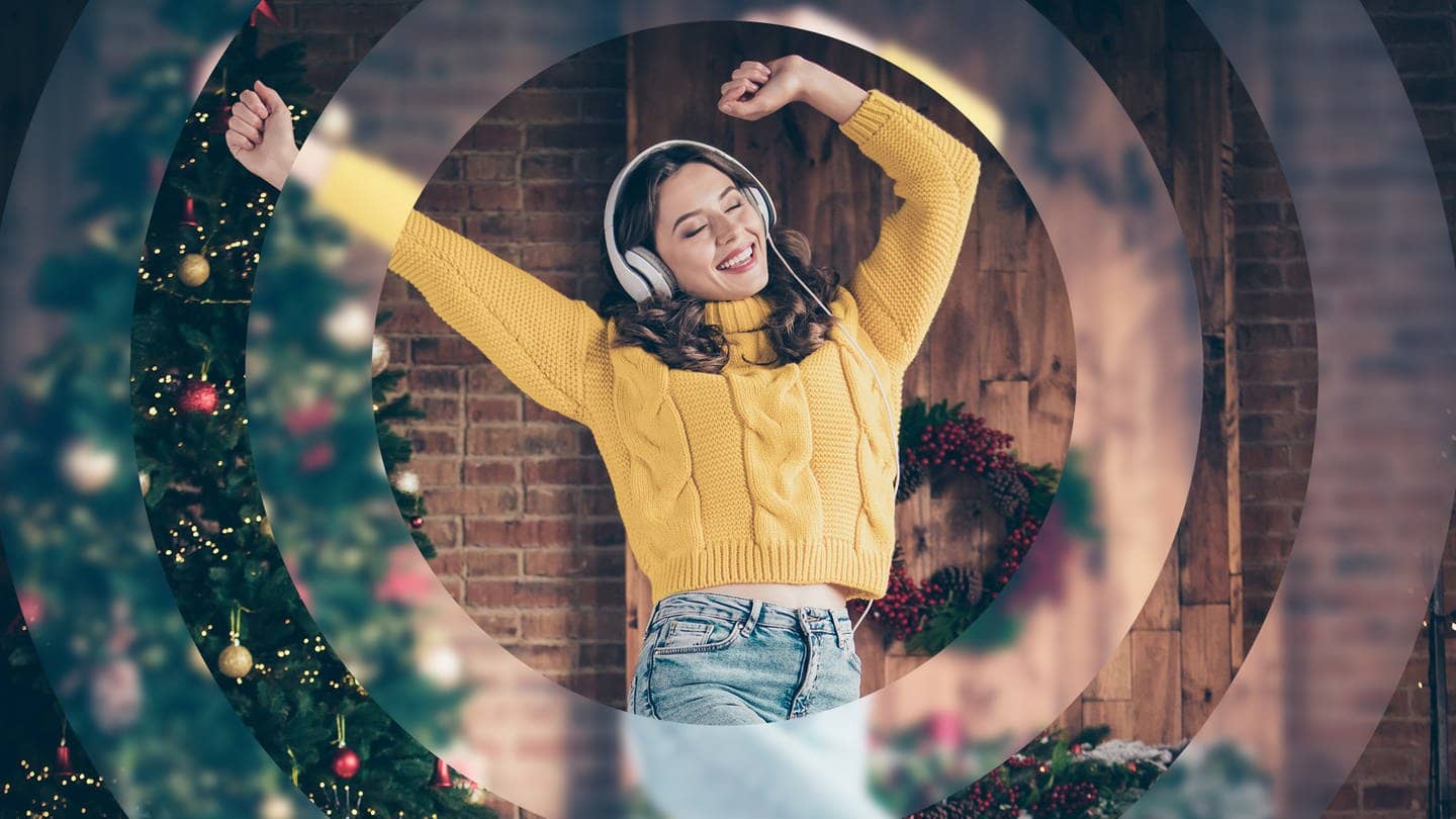 Eine junge Frau in einem gelben Pullover tanzt vor einem Weihnachtsbaum und hört Musik mit Kopfhörern.