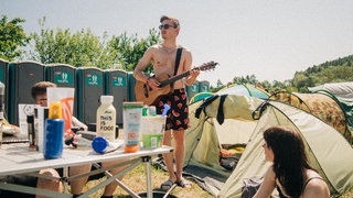 Ein junger Mann steht in Shorts und mit Gitarre auf dem Campingplatz von Rock am Ring zwischen Zelten (Foto: SWR3, SWR3 / Niko Neithardt) Ein junger Mann steht in Shorts und mit Gitarre auf dem Campingplatz von Rock am Ring zwischen Zelten