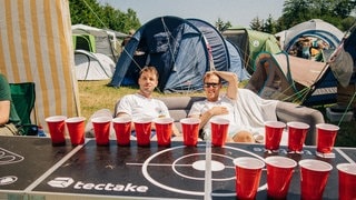 Zwei Männer sitzen hinter einem Bier-Pong-Tisch auf einem Sofa bei Rock am Ring auf dem Campingplatz (Foto: SWR3, SWR3 / Niko Neithardt) Zwei Männer sitzen hinter einem Bier-Pong-Tisch auf einem Sofa bei Rock am Ring auf dem Campingplatz