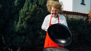 Starkoch Johann Lafer mit einem Strohballen auf dem Kopf und vor einem Grill. (Foto: SWR) Starkoch Johann Lafer mit einem Strohballen auf dem Kopf und vor einem Grill.